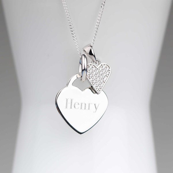 A silver heart pendant necklace with the name 'Henry' engraved on the front, hanging from a delicate silver chain. A smaller, sparkling heart charm dangles beside it against a soft white display background.