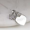 A silver heart and star pendant necklace on a delicate chain, resting on a light marble surface. The heart pendant is engraved with the name 'poppy' and is paired with a small star charm paved with clear stones.