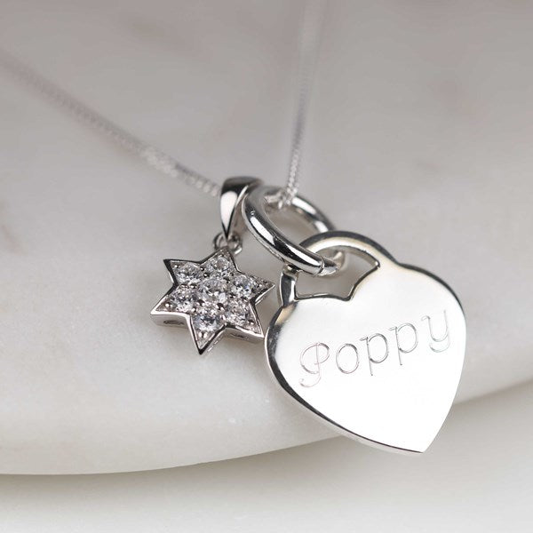 A silver heart and star pendant necklace on a delicate chain, resting on a light marble surface. The heart pendant is engraved with the name 'poppy' and is paired with a small star charm paved with clear stones.
