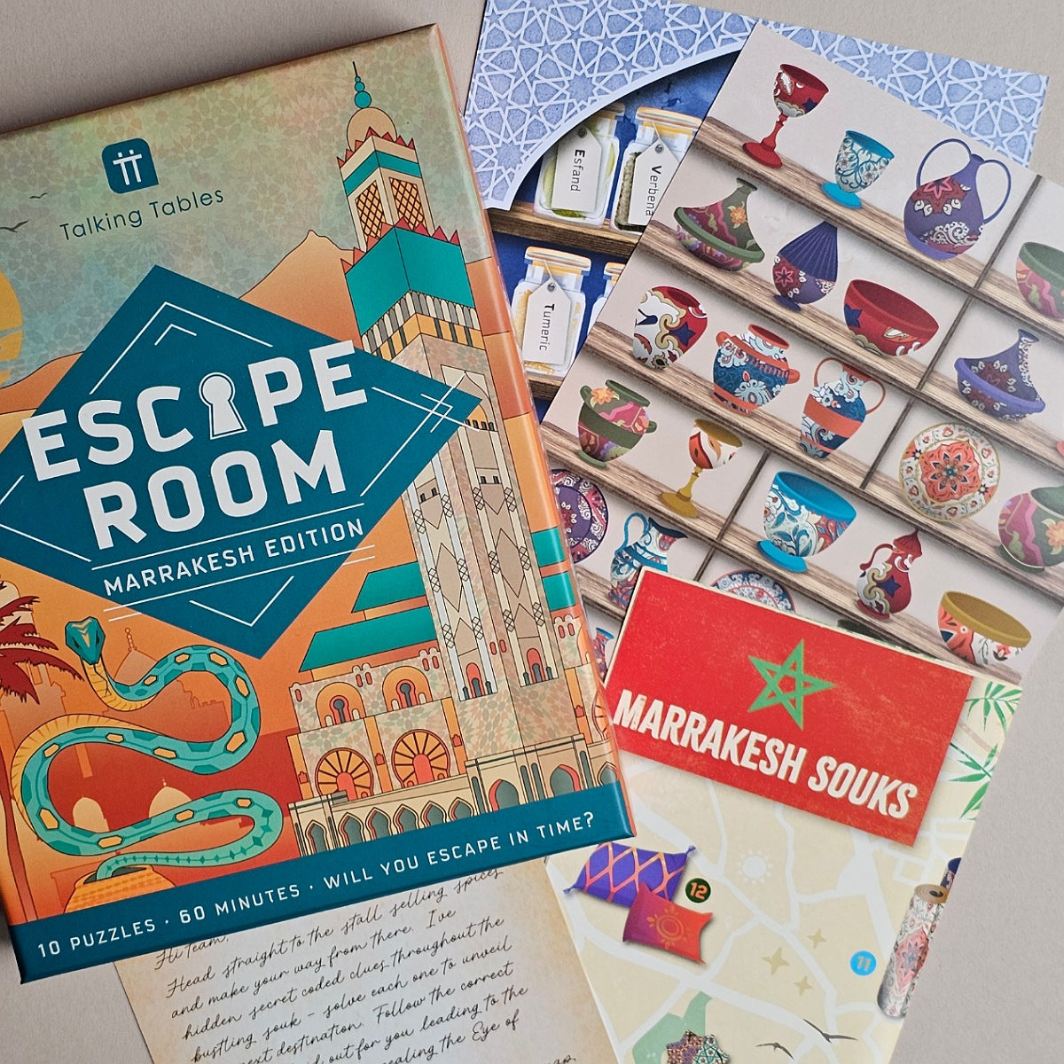 A boxed puzzle game set titled "Escape Room: Marrakesh Edition" shown with printed cards, a red "Marrakesh Souks" card, and a tray of illustrated tiles on a tabletop.