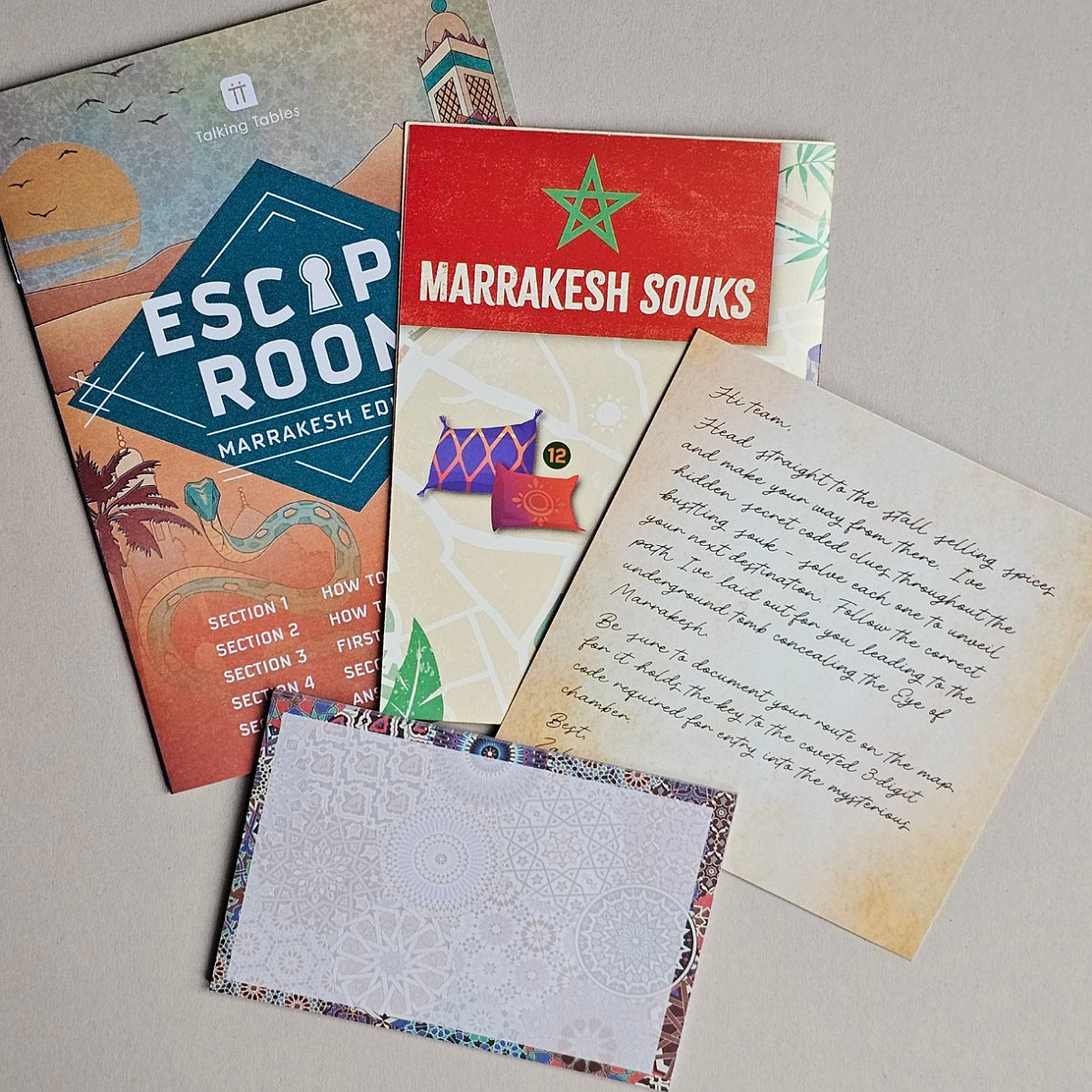 A flat lay of a travel-themed card deck or kit on a light beige surface. The set includes a colorful 'ESCAPE ROOM' booklet, a red card with 'MARRAKESH SOUKS' and a green star, a cream-colored instruction or letter card with handwritten text, a patterned envelope, and a white patterned card.