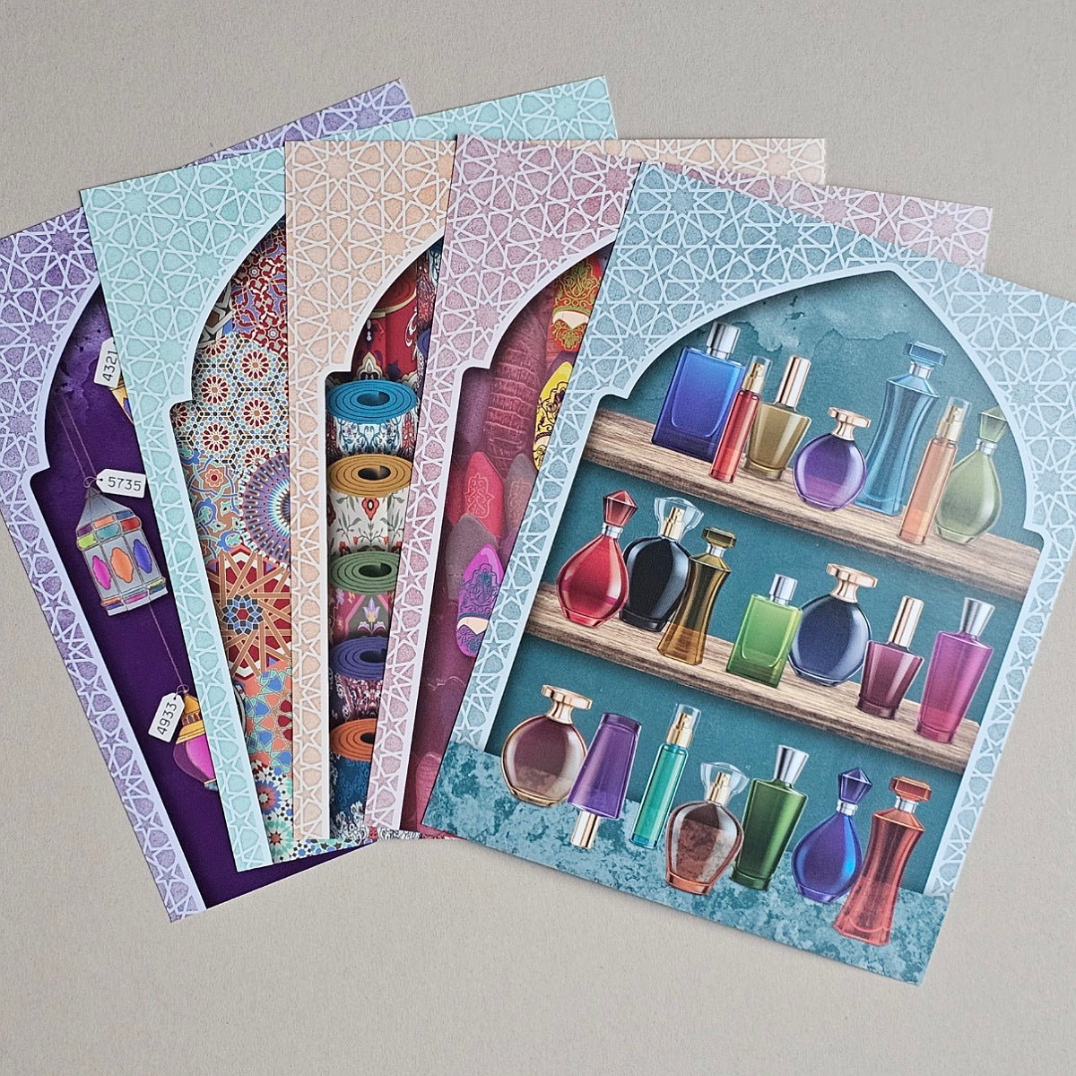A fan of colorful patterned envelopes or greeting cards arranged on a light gray surface. The front card shows an illustration of perfume bottles on shelves against a teal background, with additional patterned cards visible behind it in purple, blue, pink, and orange tones.