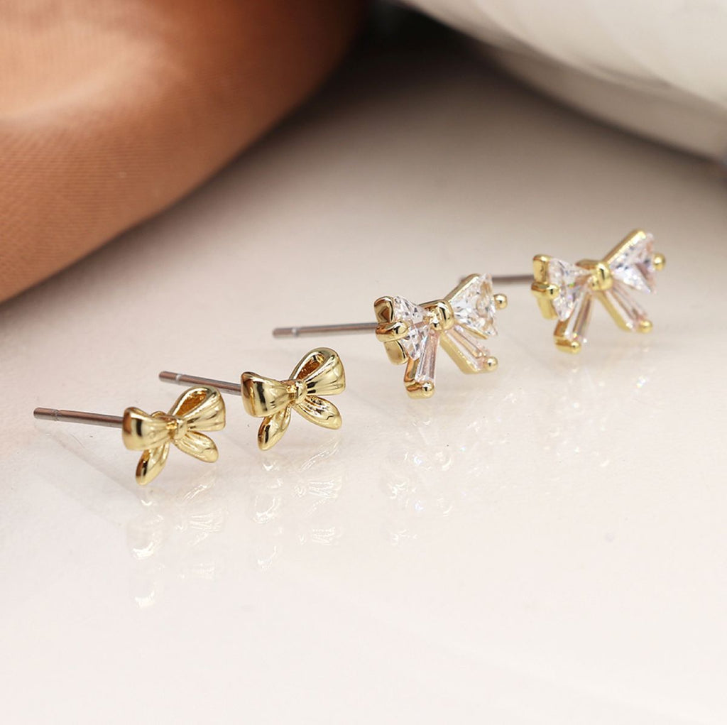Two sets of small bow-shaped stud earrings arranged diagonally on a glossy white surface. Two earrings are gold-toned with clear faceted stones, and two are gold-toned with clear faceted stones.