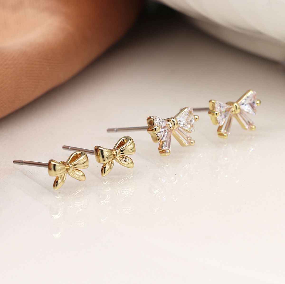 Two sets of small bow-shaped stud earrings arranged diagonally on a glossy white surface. Two earrings are gold-toned with clear faceted stones, and two are gold-toned with clear faceted stones.