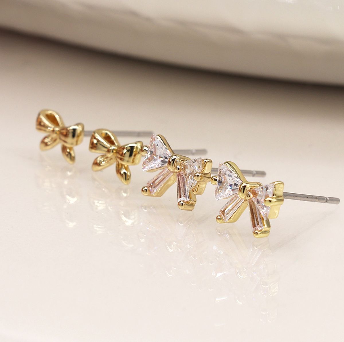 Two sets of small bow-shaped stud earrings arranged diagonally on a glossy white surface. Two earrings are gold-toned with clear faceted stones, and two are gold-toned with clear faceted stones.