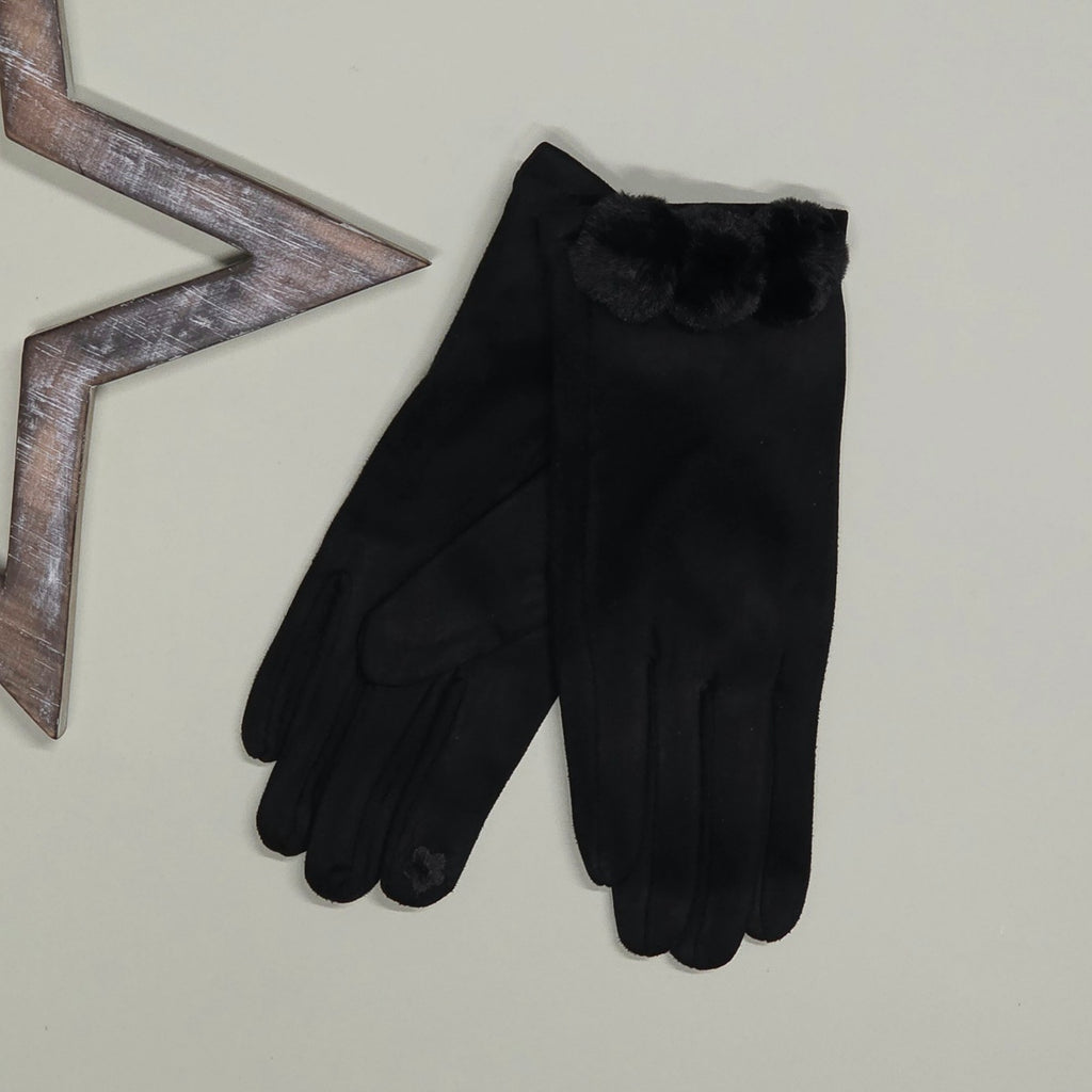 A pair of faux suede black gloves with a fuzzy black pom pom trim at the cuff, displayed on a light beige background next to a decorative wooden star prop
