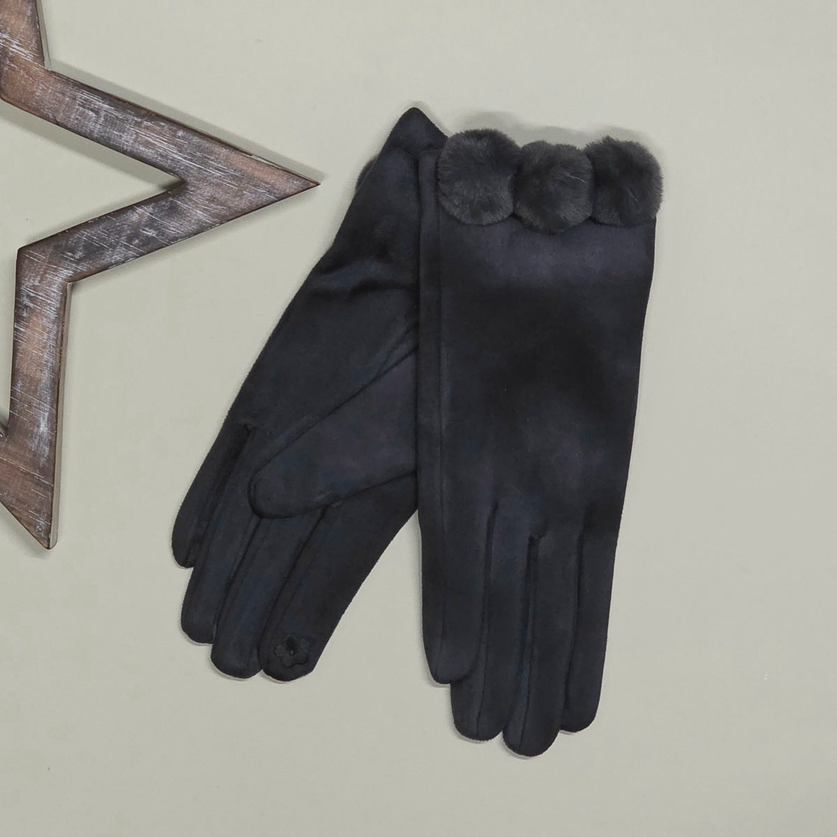 A pair of faux suede grey gloves with a fuzzy grey pom pom trim at the cuff, displayed on a light beige background next to a decorative wooden star prop