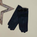 A pair of faux suede navy blue gloves with a fuzzy navy blue pom pom trim at the cuff, displayed on a light beige background next to a decorative wooden star prop