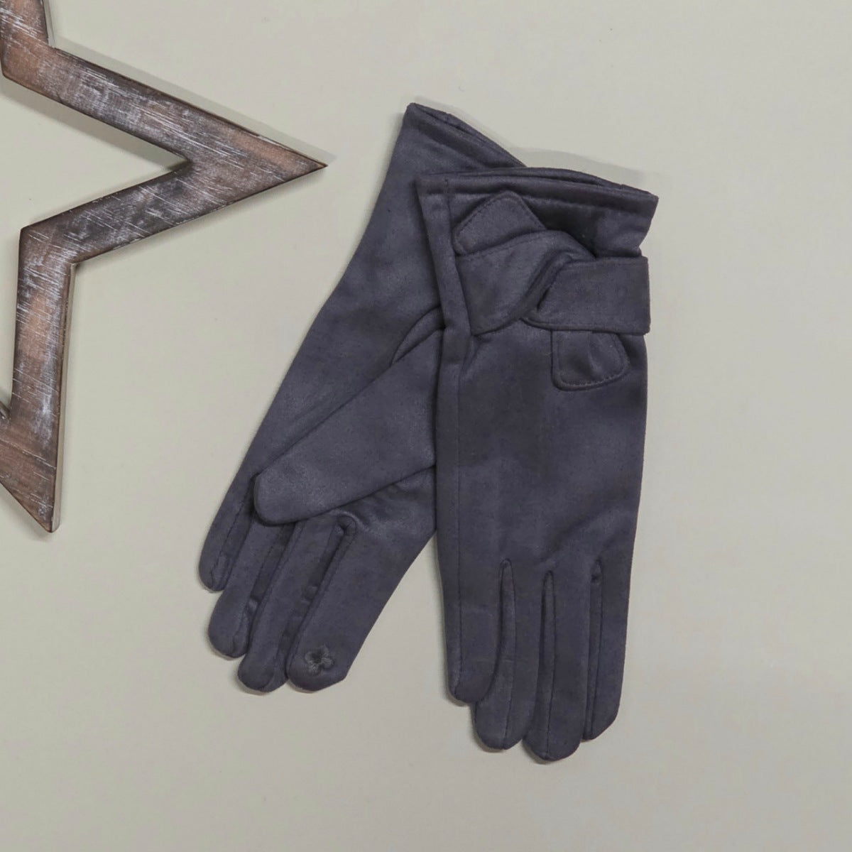 Pair of faux suede grey gloves with a twisted fabric design on a light grey surface with a decorative wooden star frame in the background.
