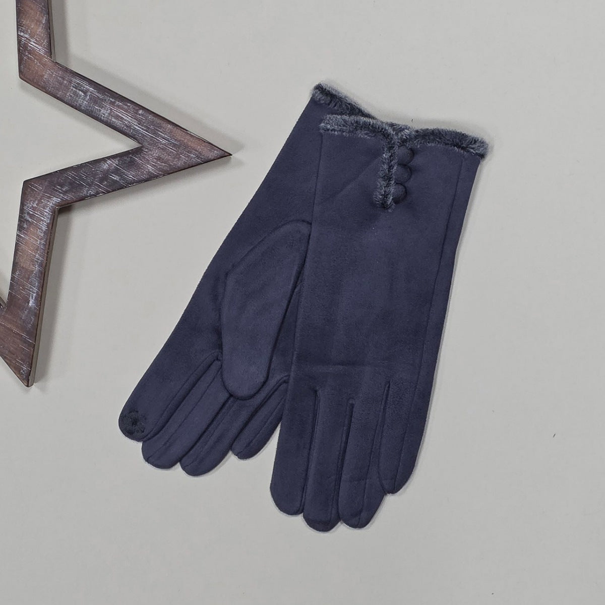 Pair of faux suede dark grey gloves with a thin grey fur trim and three dark grey buttons on a light grey surface with a decorative wooden star frame in the background.