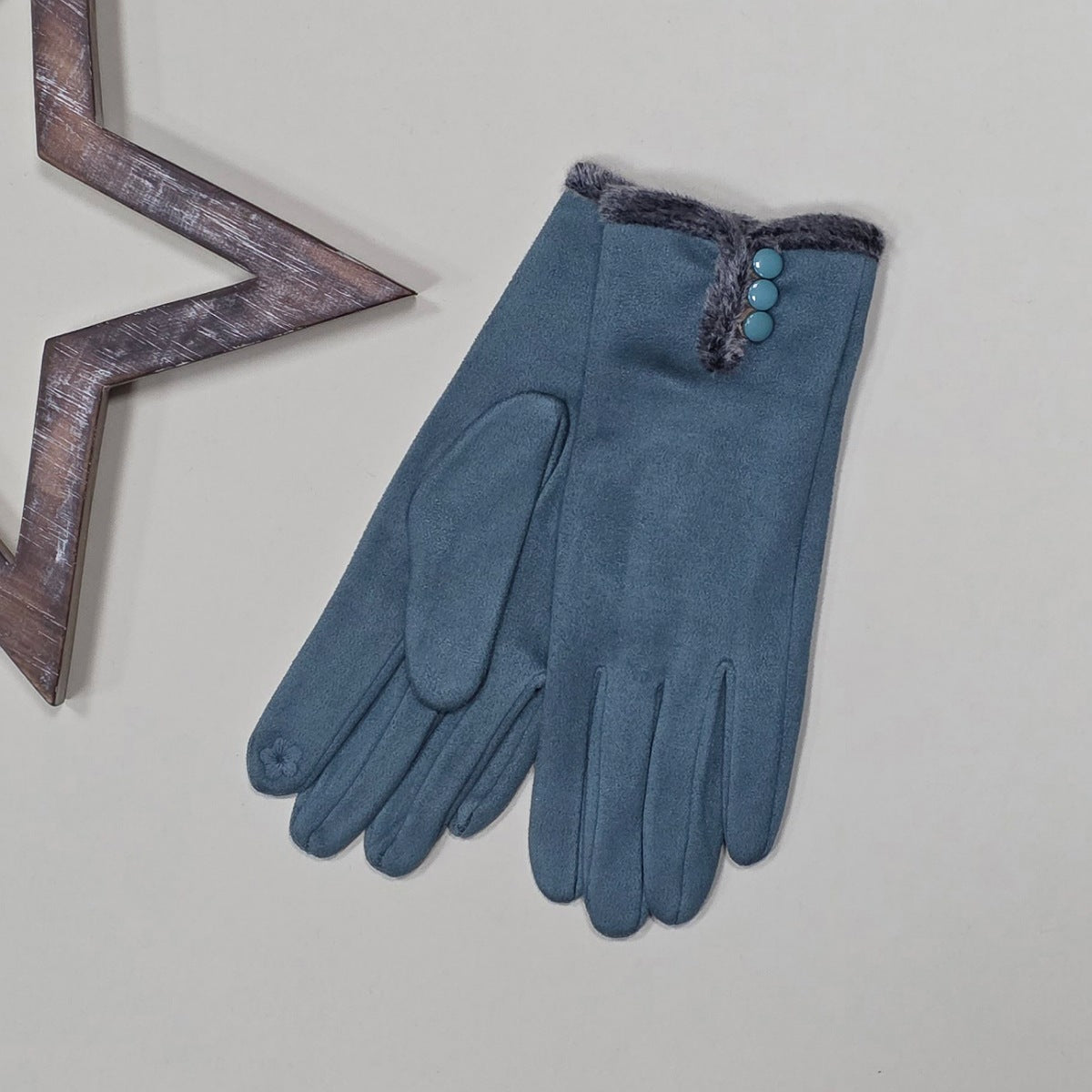 Pair of faux suede duck egg blue gloves with a thin grey fur trim and three duck egg blue buttons on a light grey surface with a decorative wooden star frame in the background.