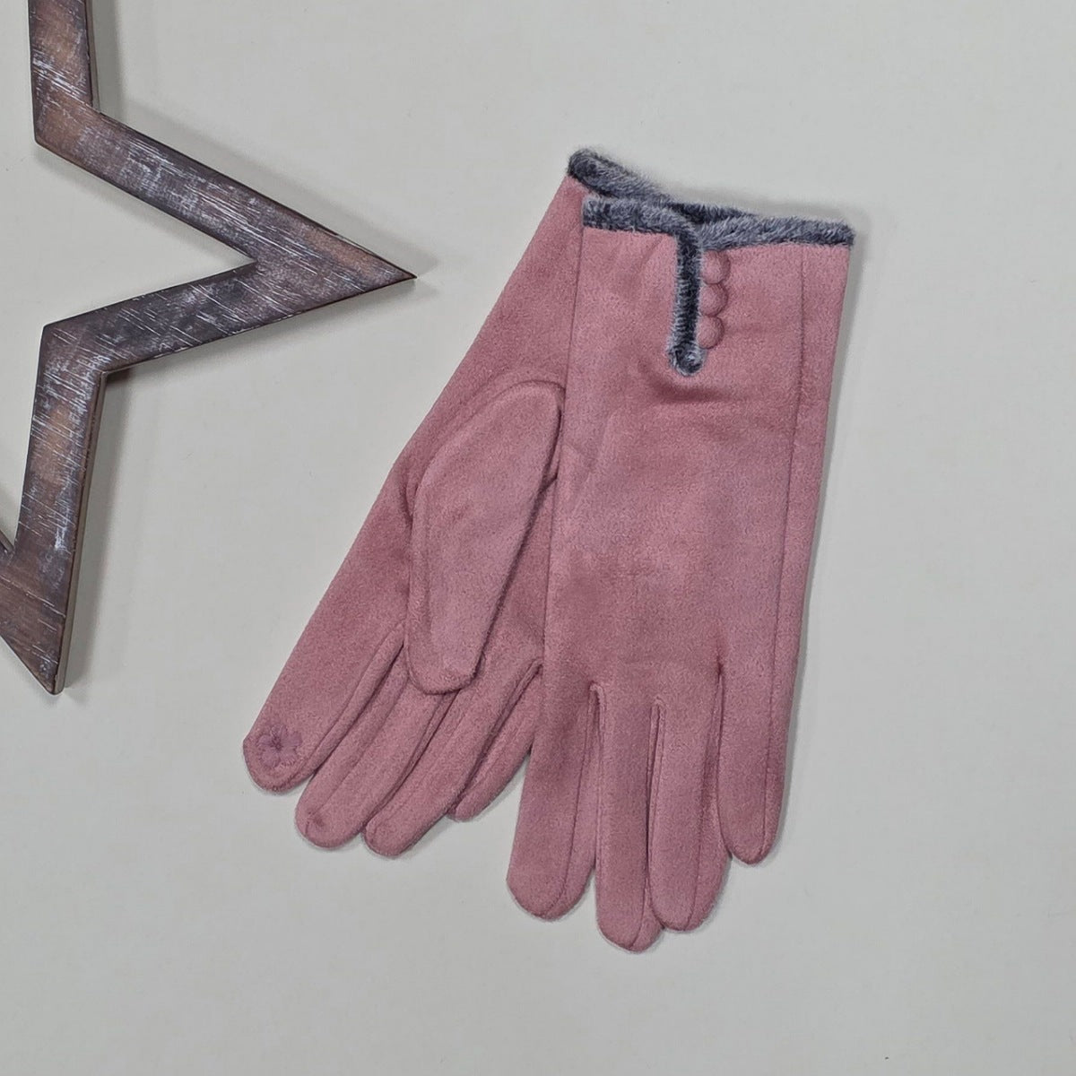 Pair of faux suede dusky pink gloves with a thin grey fur trim and three dusky pink buttons on a light grey surface with a decorative wooden star frame in the background.