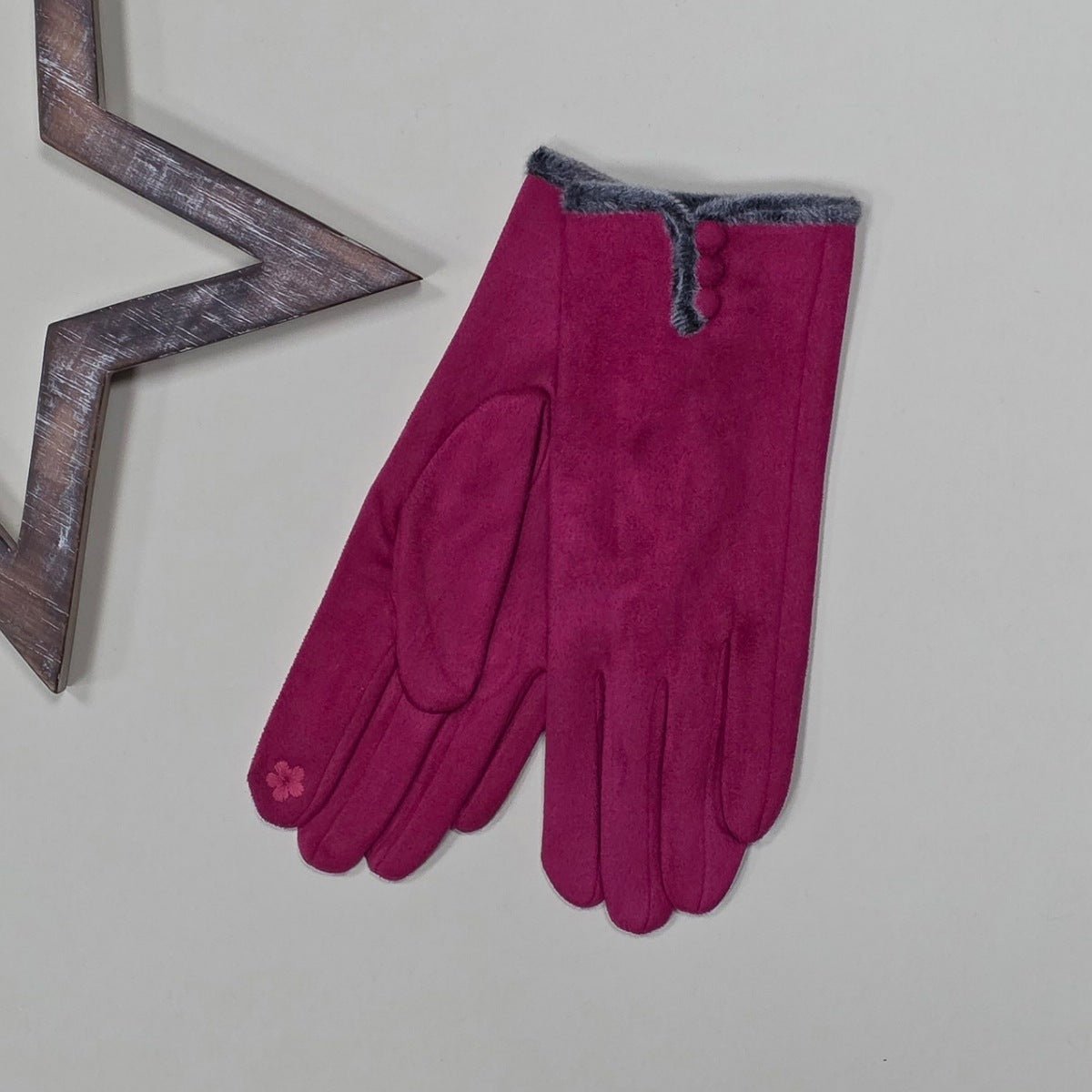 Pair of faux suede bright pink gloves with a thin grey fur trim and three bright pink buttons on a light grey surface with a decorative wooden star frame in the background.