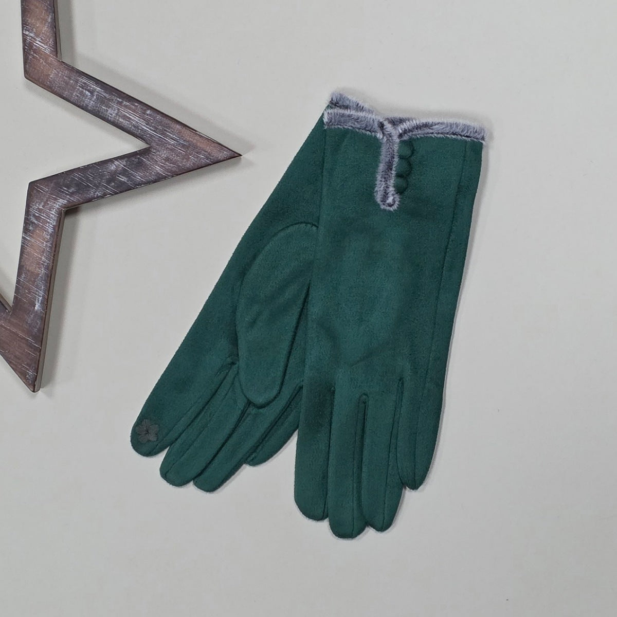 Pair of faux suede green gloves with a thin grey fur trim and three green buttons on a light grey surface with a decorative metal frame in the background.