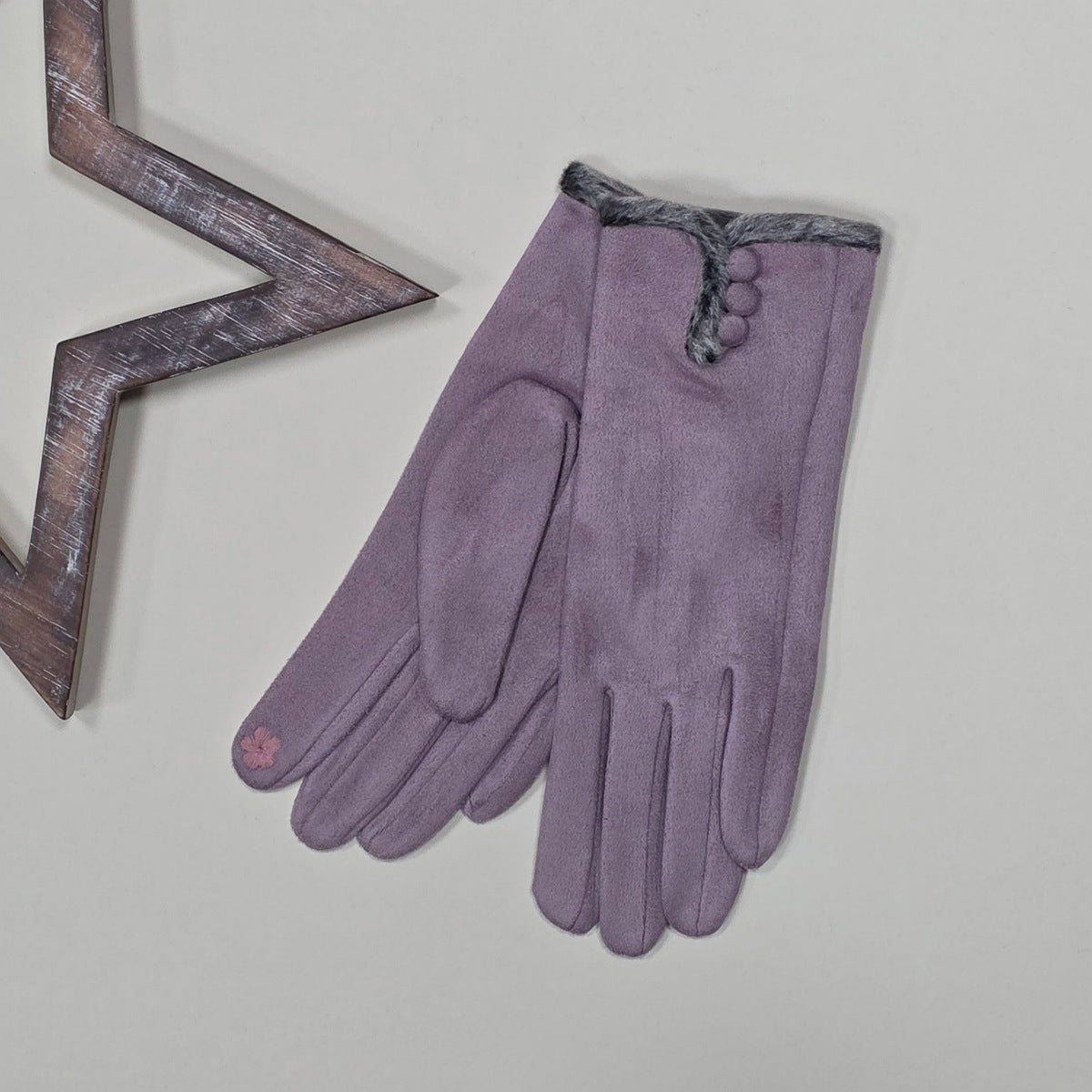 Pair of faux suede mauve gloves with a thin grey fur trim and three mauve buttons on a light grey surface with a decorative metal frame in the background.