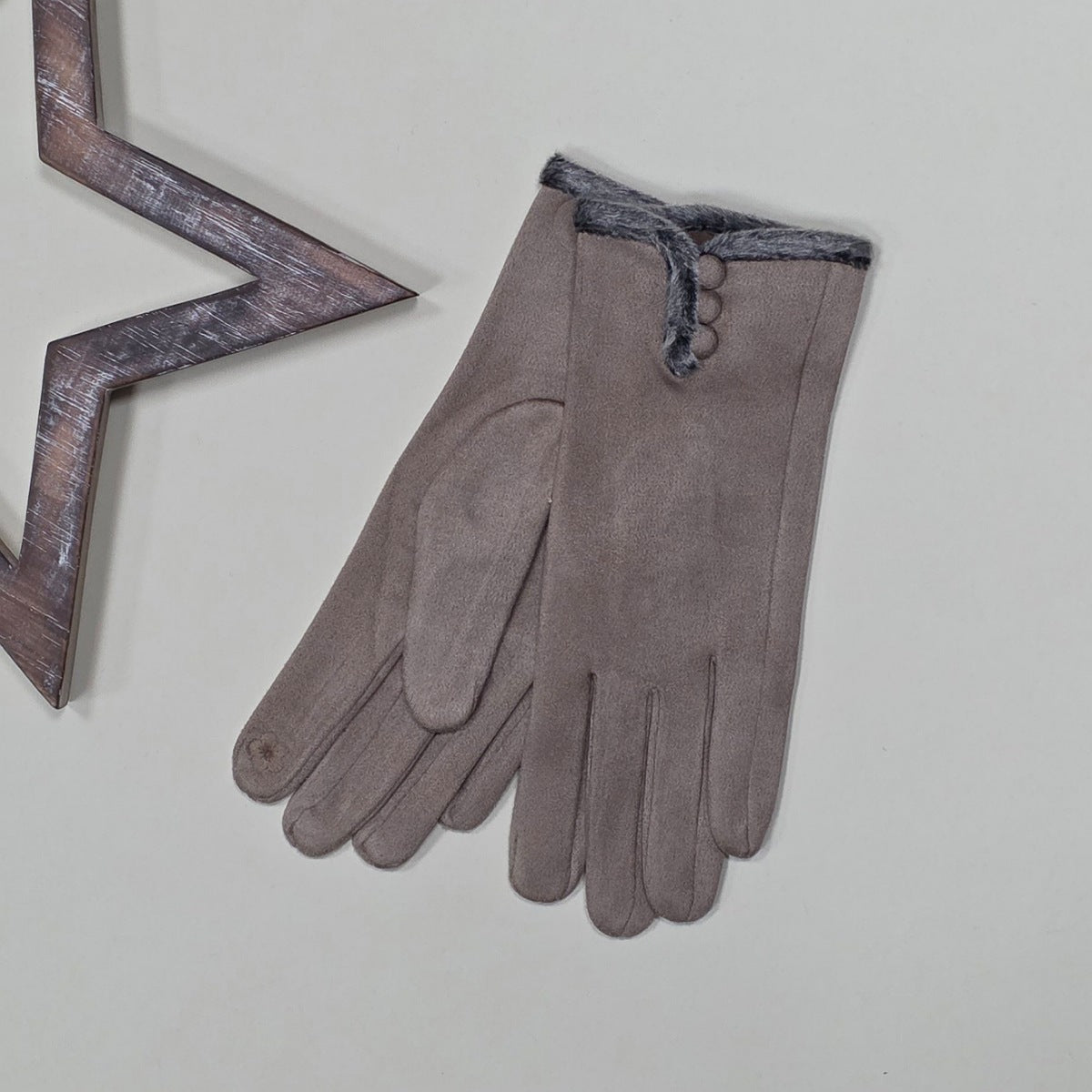 Pair of faux suede mocha gloves with a thin grey fur trim and three mocha buttons on a light grey surface with a decorative metal frame in the background.