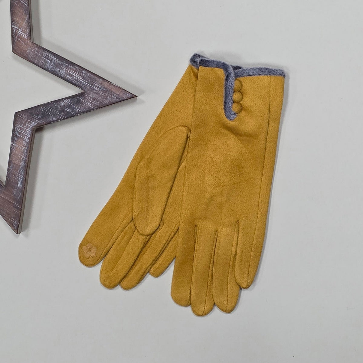 Pair of faux suede mustard gloves with a thin grey fur trim and three mustard buttons on a light grey surface with a decorative metal frame in the background.