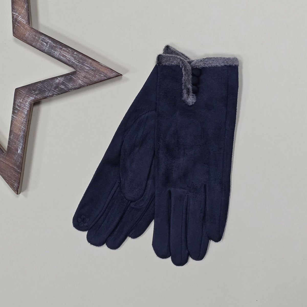 Pair of faux suede navy gloves with a thin grey fur trim and three navy buttons on a light grey surface with a decorative metal frame in the background.