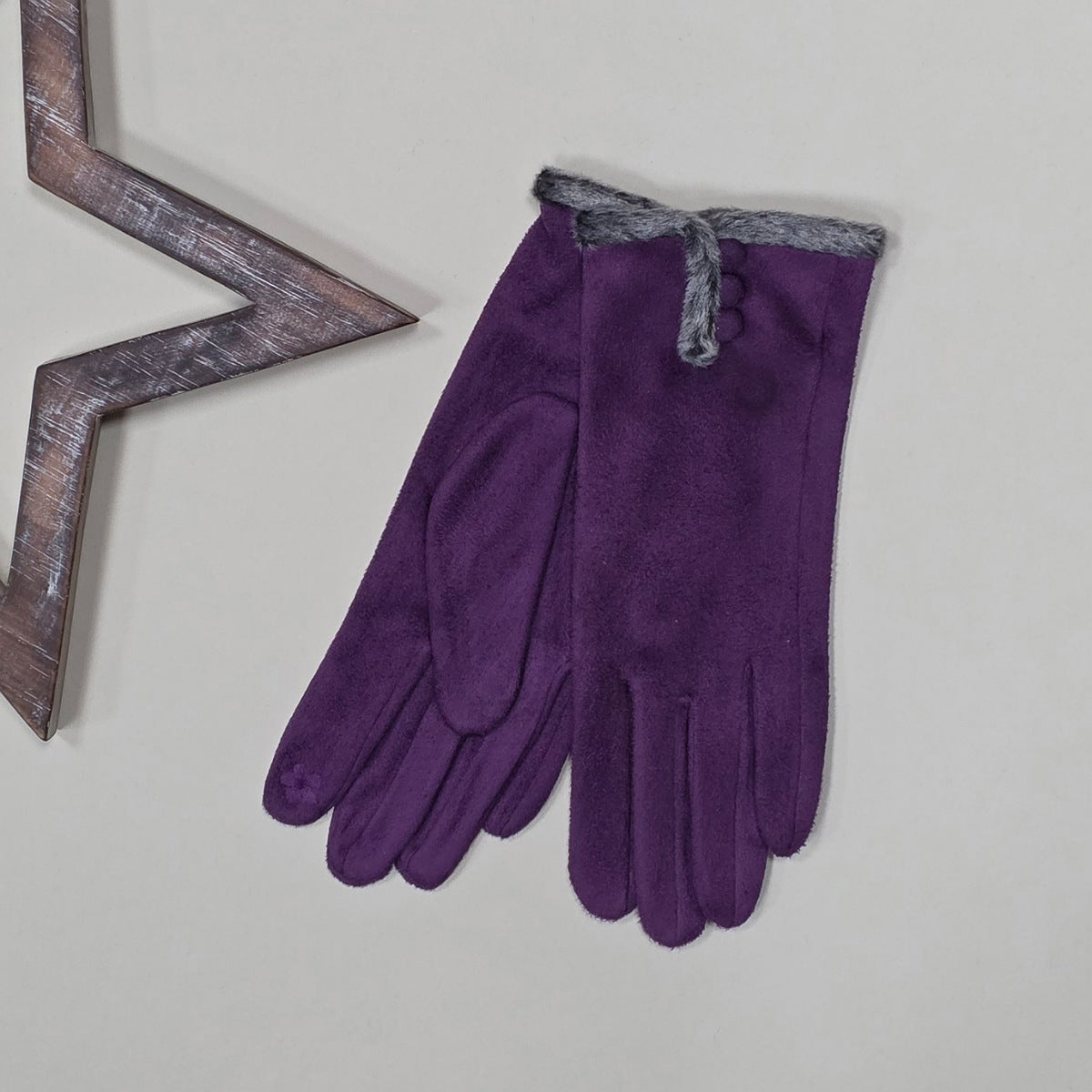 Pair of faux suede purple gloves with a thin grey fur trim and three purple buttons on a light grey surface with a decorative metal frame in the background.
