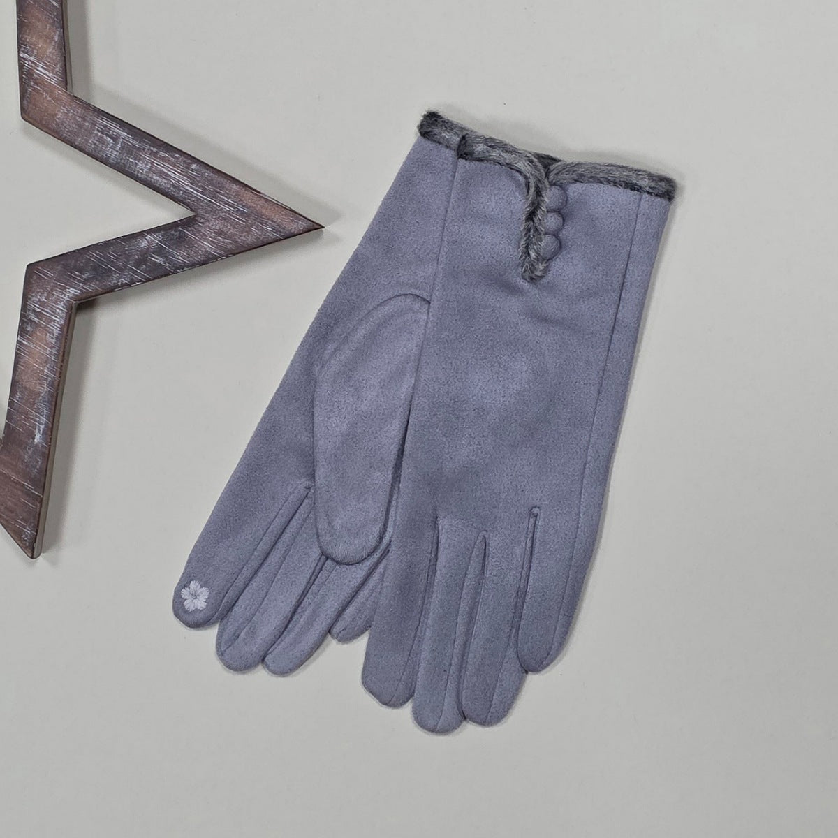 Pair of faux suede silver gloves with a thin grey fur trim and three silver buttons on a light grey surface with a decorative metal frame in the background.