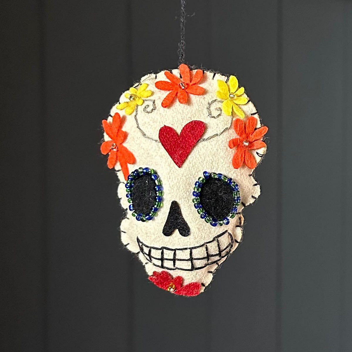 A handmade felt sugar skull ornament hanging against a dark grey panelled background. The skull is off-white felt decorated with a red felt heart on the forehead, orange and yellow felt flowers, black felt eye sockets with blue bead trim, and a red felt mouth detail.