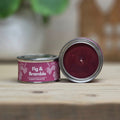 A small round metal tin candle labeled 'Fig & Bramble Scent Palette' sits on a wooden surface with its lid removed, showing a deep burgundy wax inside. The background is softly blurred with green leaves and warm wooden decor.