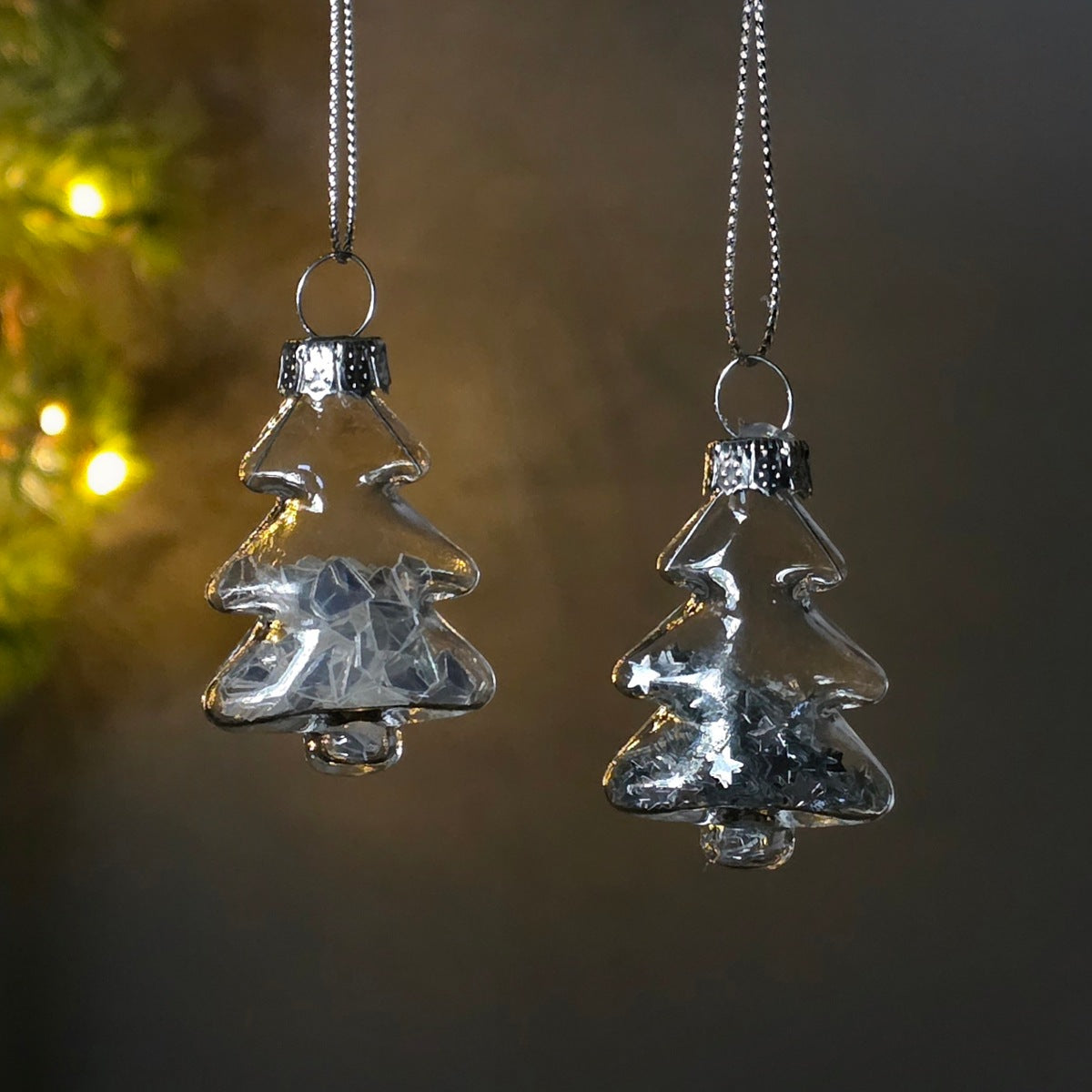 Two clear glass Christmas tree ornaments with silver sequin shapes inside with silver caps and hanging silver chains, shown against a dark blurred background with warm lights.
