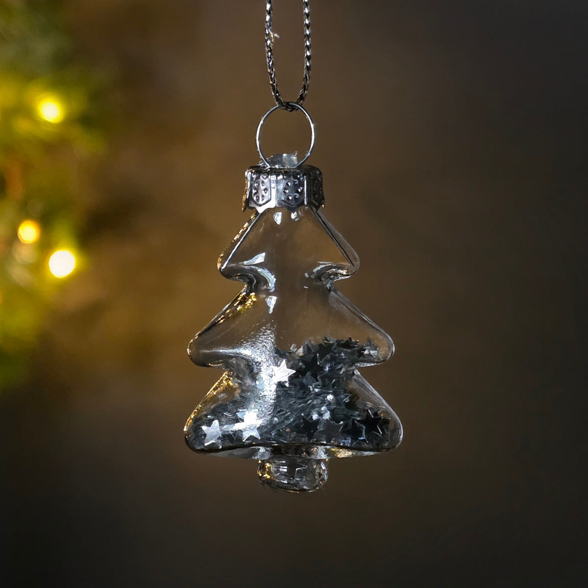 A clear glass Christmas tree ornament hanging from a silver chain, filled with silver glitter and small star-shaped pieces, set against a dark background with soft warm bokeh lights.