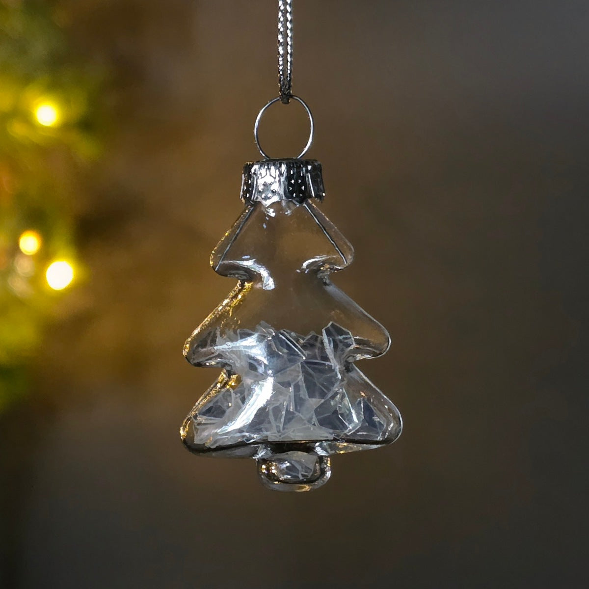 A clear glass Christmas tree ornament containing geometric shapes, hanging from a silver metal ring against a dark, blurred background with warm bokeh lights.