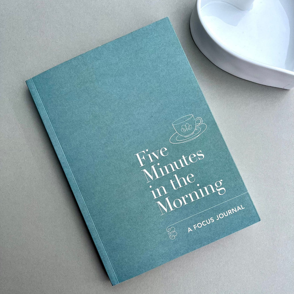 A teal blue hardcover journal titled 'Five Minutes in the Morning' by A FOCUS JOURNAL, placed diagonally on a neutral grey surface next to a white ceramic dish.
