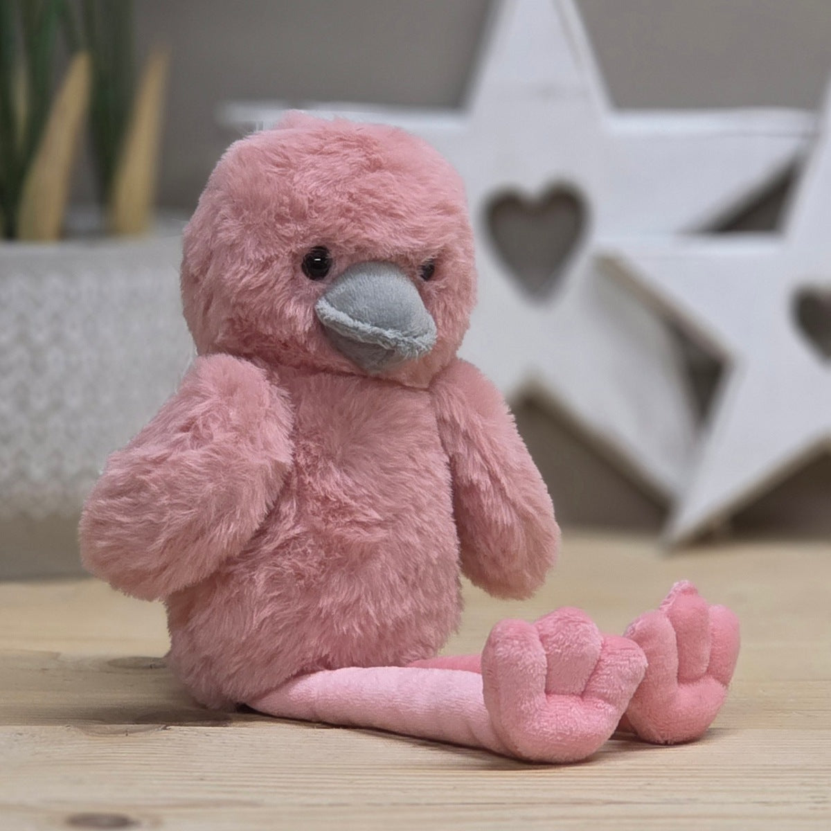 Pink plush soft fuzzy flamingo toy with long legs, grey beak and black eyes, sat on a wooden surface with a blurred natural background