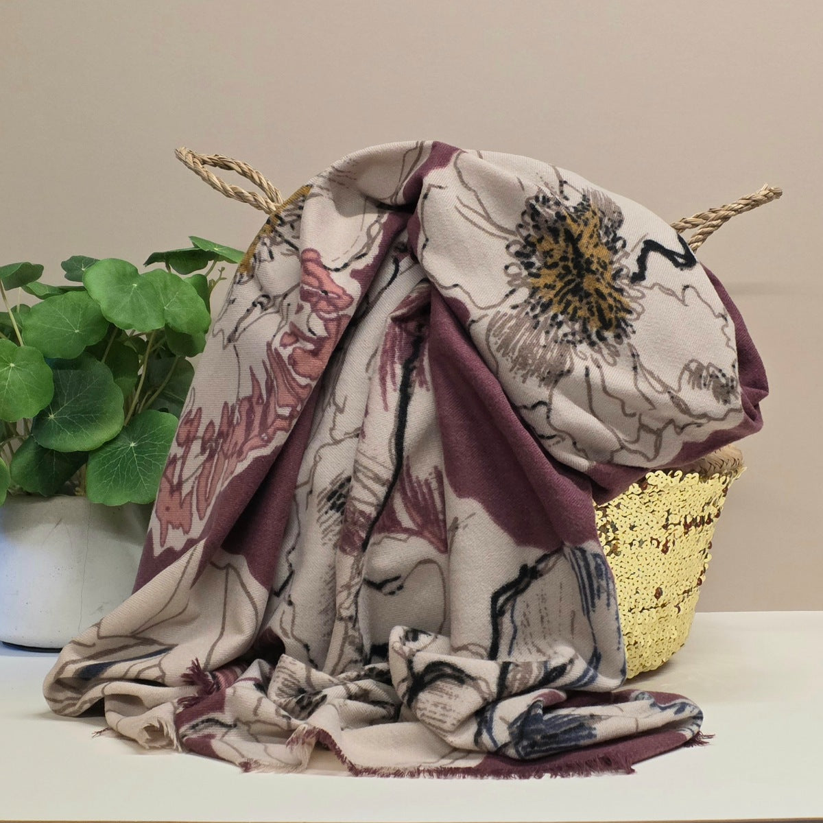 A mid weight patterned scarf draped over a woven basket, with a potted green plant on the left and a gold sequined tray on the right, set against a plain beige wall.