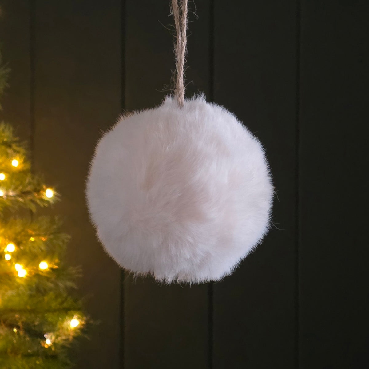 A fluffy white faux-fur ball shaped hanging ornament with a natural jute string, shown against a dark panelled background with warm yellow Christmas lights on the left.