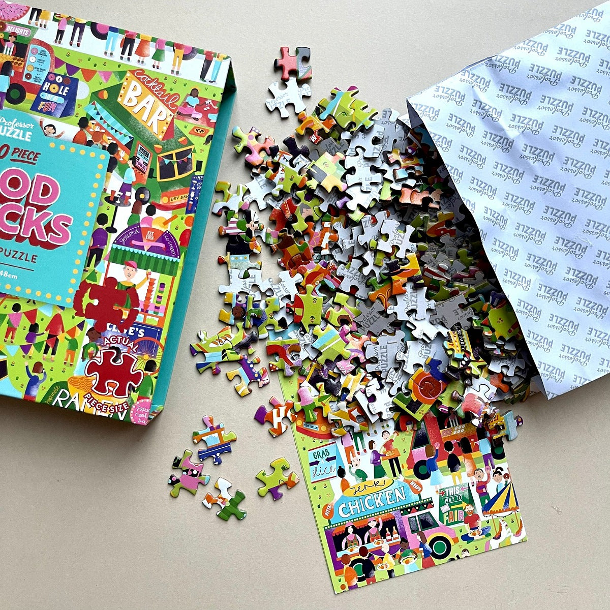 A colorful jigsaw puzzle box shown on a beige surface with many loose puzzle pieces spilled out and one completed puzzle piece cluster displayed. The box and pieces feature a busy illustrated design.