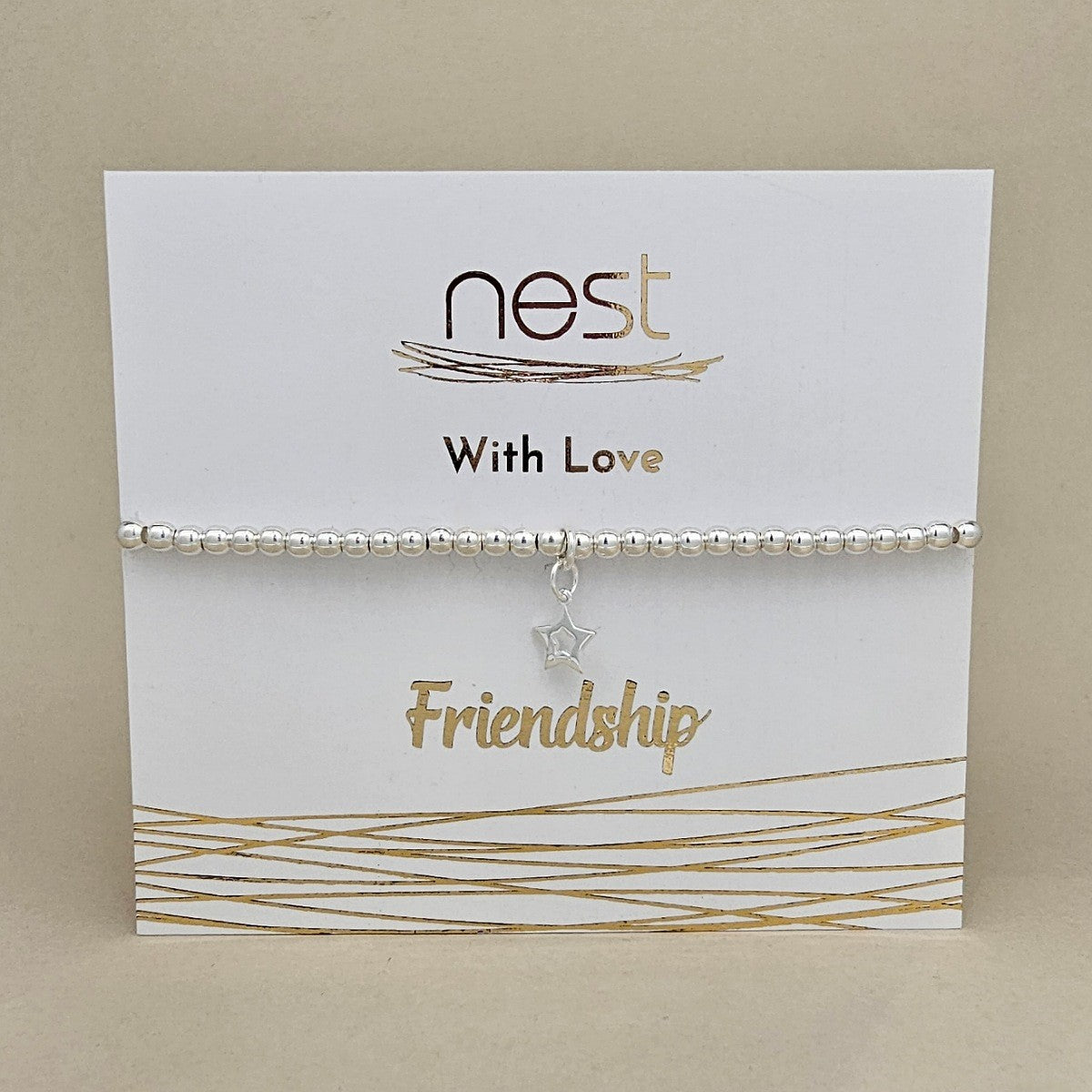 A silver beaded bracelet with a dangling star charm, presented on a white display card with gold foil text reading 'nest With Love Friendship'.