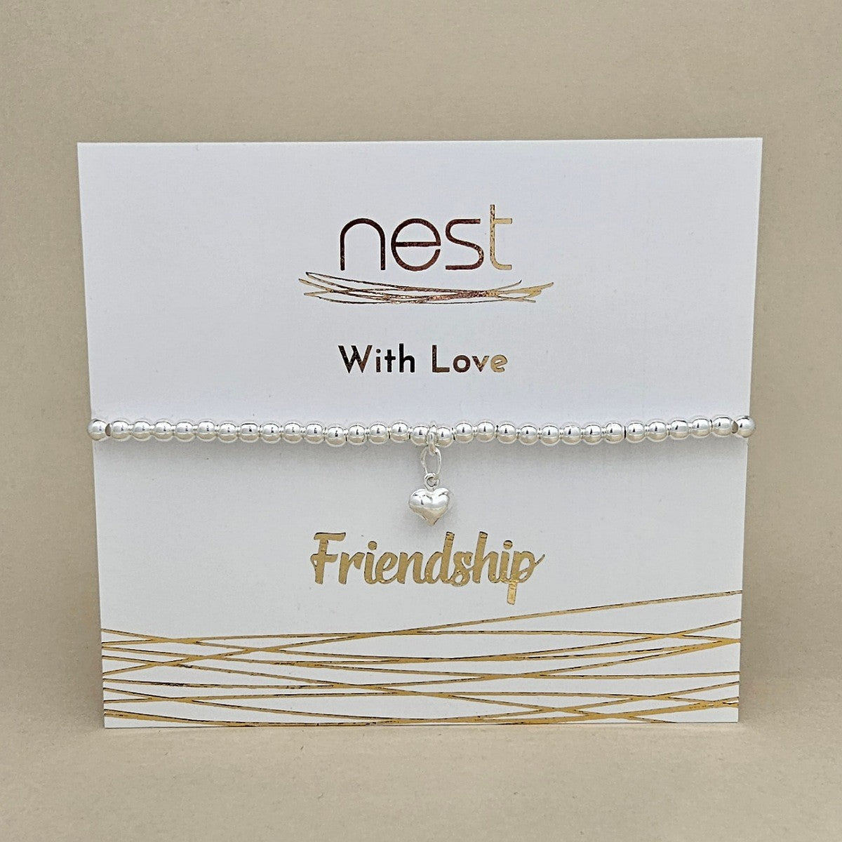 A silver beaded bracelet with a small dangling heart charm, displayed on a white card that reads 'nest With Love Friendship' with gold decorative lines, set against a beige background.