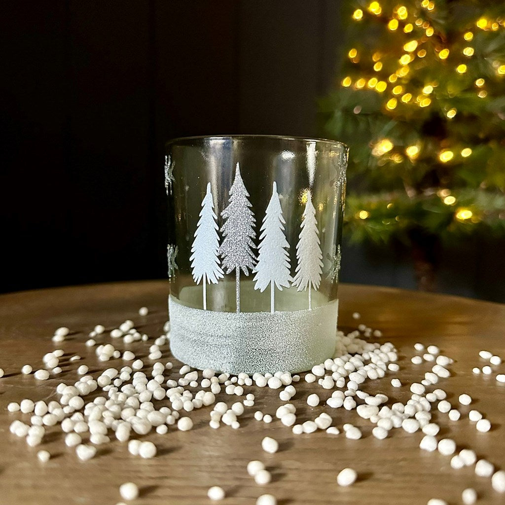 Decorative glass tea light holder with white and silver tree etching, displayed on a wooden surface with white beads and a blurred Christmas tree in the background.
