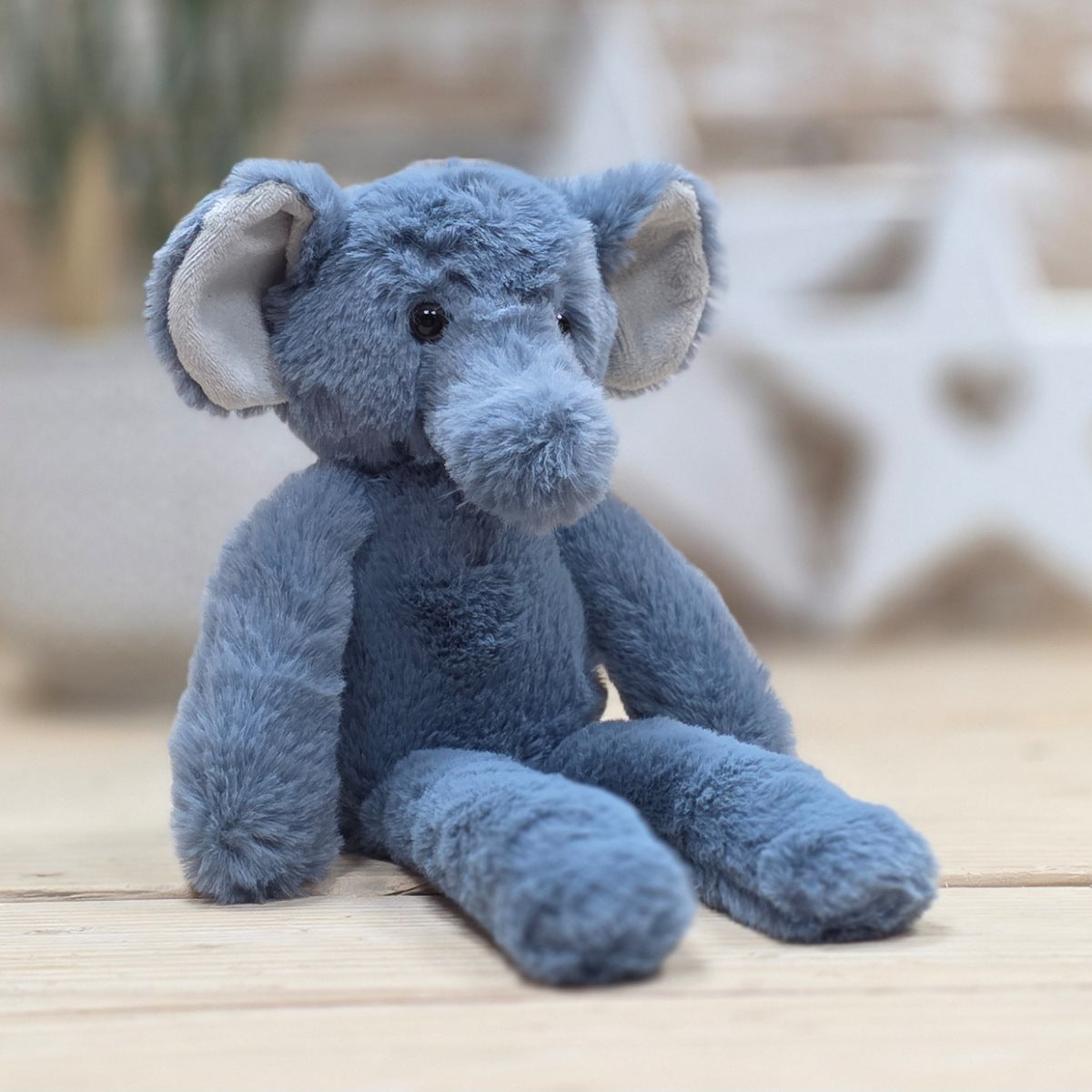 Blue plush soft fuzzy elephant toy  with grey ears and black eyes, on a wooden surface with a blurred natural background