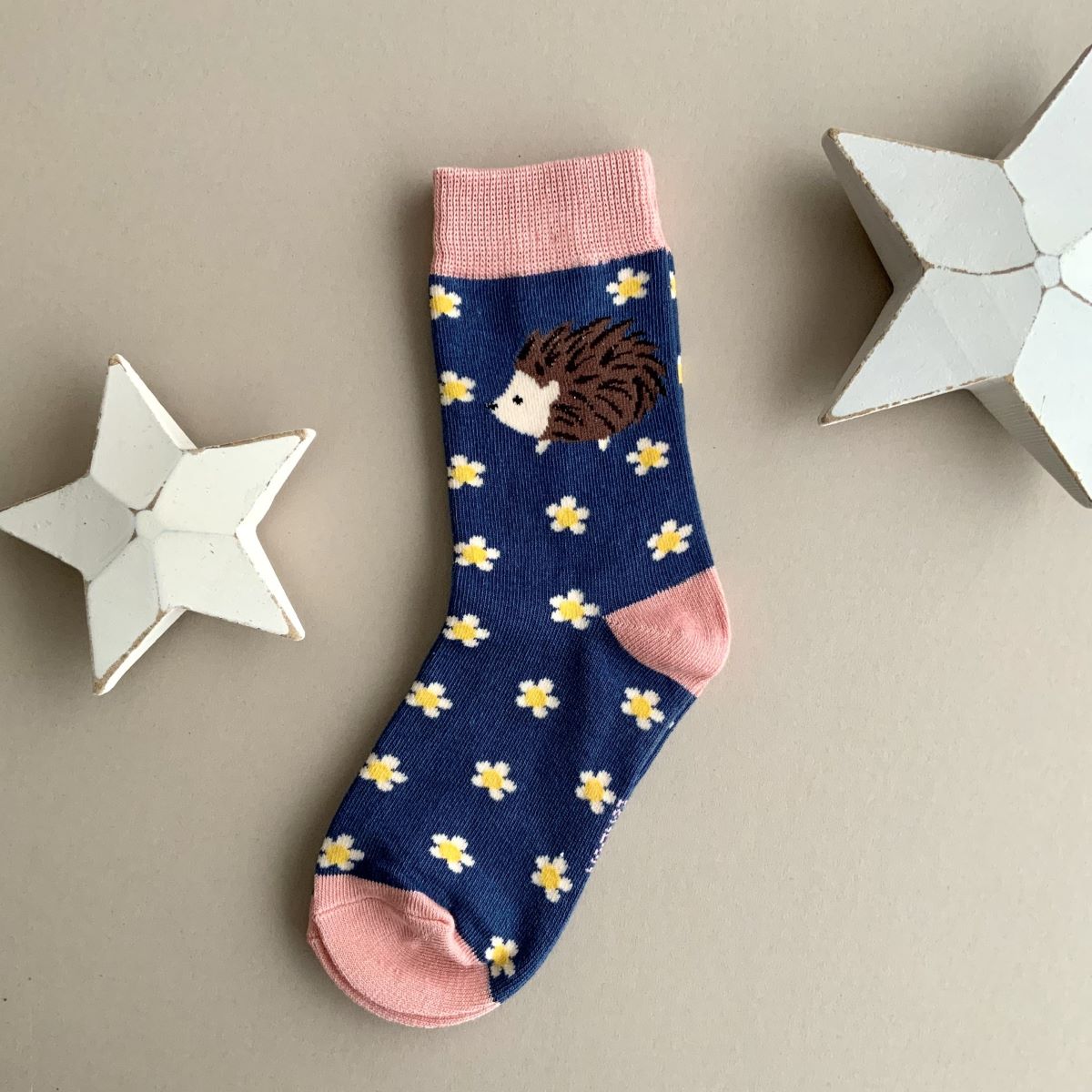 A single navy blue sock with a repeating pattern of white and yellow daisy flowers with a single brown hedgehog.  Featuring a pink cuff, heal and toe, displayed on a neutral beige background with white star-shaped props.
