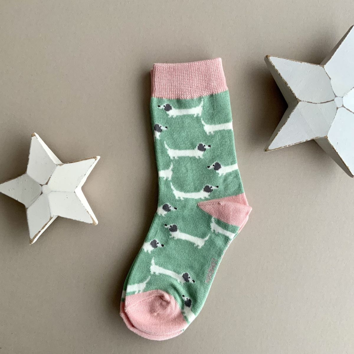 A single mint green sock with a repeating pattern of white sausage dogs with grey ears.  Featuring a pink cuff, heal and toe, displayed on a neutral beige background with white star-shaped props.

