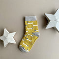 A single yellow sock with a repeating pattern of white sausage dogs with grey ears.  Featuring a light grey cuff, heal and toe, displayed on a neutral beige background with white star-shaped props.
