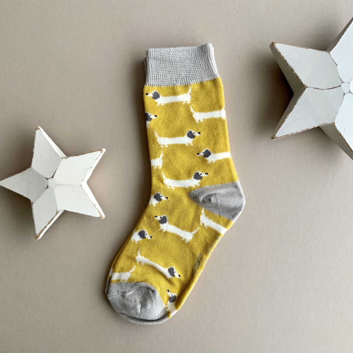 A single yellow sock with a repeating pattern of white sausage dogs with grey ears.  Featuring a light grey cuff, heal and toe, displayed on a neutral beige background with white star-shaped props.
