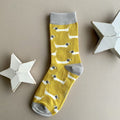 A single yellow sock with a repeating pattern of white sausage dogs with grey ears.  Featuring a light grey cuff, heal and toe, displayed on a neutral beige background with white star-shaped props.
