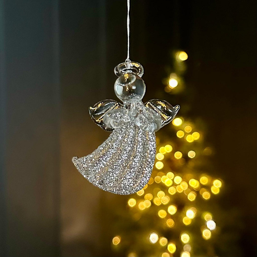 A clear glass angel Christmas ornament with a glittery silver dress and wings, hanging against a dark background with warm yellow bokeh lights.