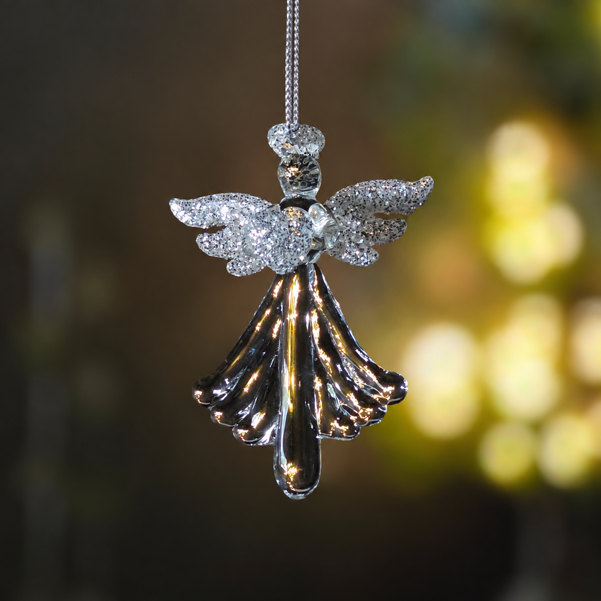 A hanging angel ornament with glittery silver wings and a shiny dark metallic dress, suspended by a thin cord against a softly blurred warm-toned background.