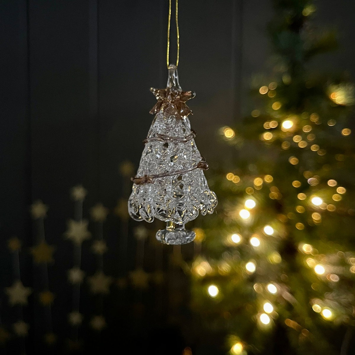 Decorative glass Christmas tree ornament with gold star and wrap around garland hanging against a blurred Christmas tree background.