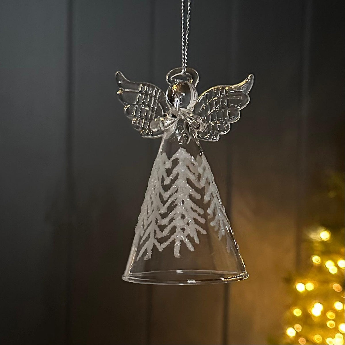 A clear glass angel Christmas ornament with a bell-shaped body, detailed wings, and a glittery chevron pattern, hanging against a dark wooden background with warm bokeh lights.