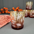 A frosted glass candle holder with a warm amber-brown tint and white botanical leaf pattern, styled on a grey surface with a striped cloth and dried orange flowers in the background.