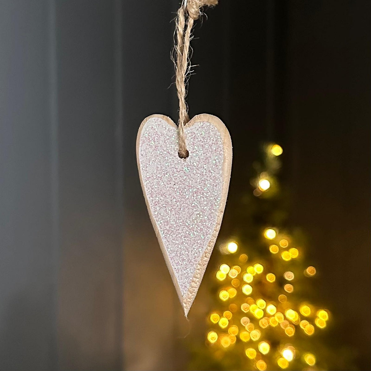 A glittery light pink heart-shaped wooden hanging ornament suspended by a natural twine string, with a warm Christmas tree light in the background.