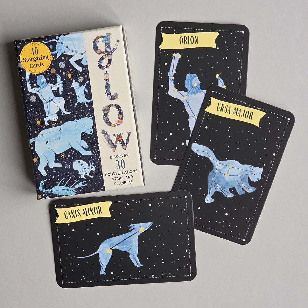 A boxed card deck and three zodiac cards arranged on a light grey surface. The box reads 'Glow' and says it contains 30 constellations cards.